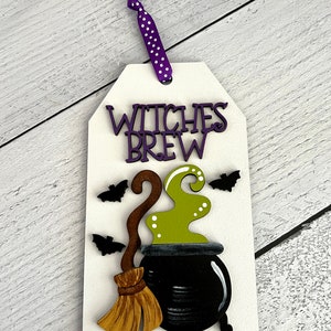 May include: A white wooden sign with the text "Witches Brew" in purple letters. The sign features a black cauldron with green smoke, a brown broom, and black bats.