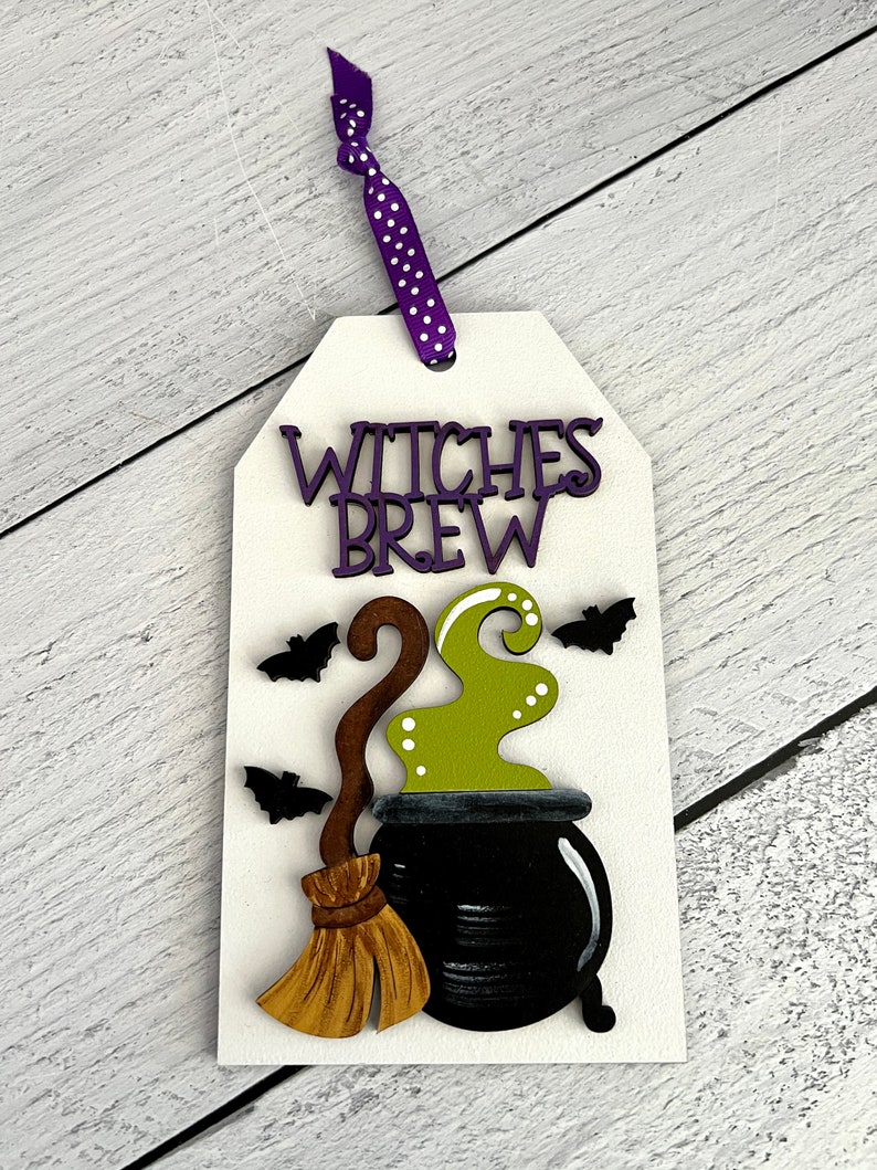 May include: A white wooden sign with the text "Witches Brew" in purple letters. The sign features a black cauldron with green smoke, a brown broom, and black bats.