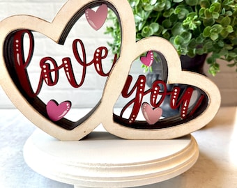 May include: A wooden heart-shaped decoration with the words "Love You" in red script. The decoration features two connected hearts, each with a pink heart accent. It is displayed on a white, round pedestal.