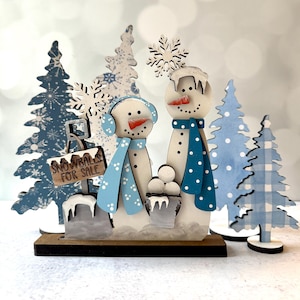 May include: Wooden winter scene featuring two snowmen with carrot noses and blue scarves, surrounded by blue snowflake trees. One snowman wears earmuffs and the other has a bucket of snowballs. A sign reads "Snowballs for Sale."