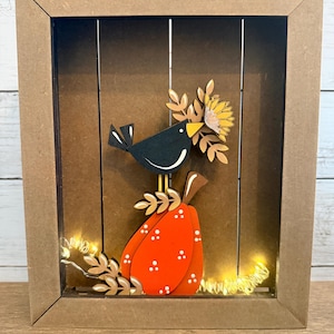 May include: A shadow box frame containing a layered wooden cutout of a black bird perched on an orange pumpkin with white polka dots. The bird holds a yellow sunflower and brown leaves in its beak. Warm string lights are tucked at the base.