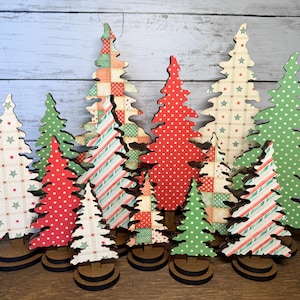 May include: A collection of twelve flat, cutout Christmas tree decorations standing on circular wooden bases. The trees feature various patterns including red and green polka dots, diagonal stripes, and star-filled grids in festive holiday colors.