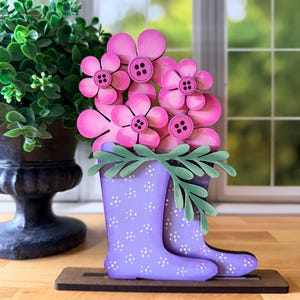 May include: A decorative wooden floral arrangement featuring pink flowers with brown button centers and green leaves, displayed in purple rain boots with white polka dots. The boots sit on a brown wooden base.