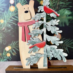 以下が含まれることがあります： 赤いスカーフをつけたクマと、2羽の赤い鳥がいるクリスマスツリーが描かれた木製のクリスマス飾りです。クマと木は薄茶色で、緑と赤のアクセントがあります。装飾は茶色のベースにあります。