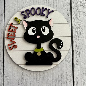 May include: A round wooden sign with a black cat and the text "Sweet & Spooky" in purple and orange letters.
