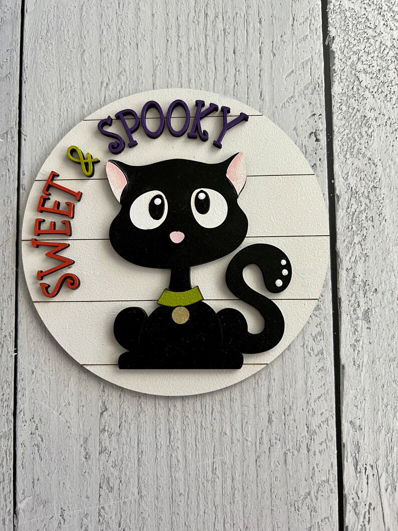 May include: A round wooden sign with a black cat and the text "Sweet & Spooky" in purple and orange letters.