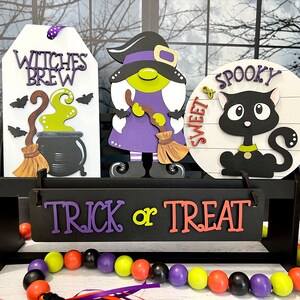 May include: A wooden shelf with three Halloween decorations: a witch with a purple dress and a broom, a sign that says "Witches Brew" with a cauldron and a broom, and a round sign that says "Sweet & Spooky" with a black cat. The shelf has a sign that says "Trick or Treat" and a beaded garland with black, purple, yellow, and orange beads.