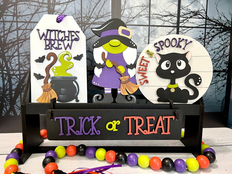 May include: A wooden shelf with three Halloween decorations: a witch with a purple dress and a broom, a sign that says "Witches Brew" with a cauldron and a broom, and a round sign that says "Sweet & Spooky" with a black cat. The shelf has a sign that says "Trick or Treat" and a beaded garland with black, purple, yellow, and orange beads.