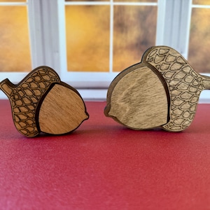 May include: Two wooden acorn ornaments, one larger than the other, with a detailed engraved pattern on the cap. The acorns are a light brown color and have a natural wood grain.