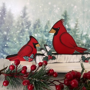 Pode incluir: Uma decora&ccedil;&atilde;o de Natal de madeira com dois cardeais vermelhos empoleirados em um galho de b&eacute;tula branco com azevinho verde e bagas vermelhas. Os cardeais s&atilde;o pintados em um estilo realista com olhos e bicos pretos. O fundo &eacute; uma imagem desfocada de uma floresta nevada.