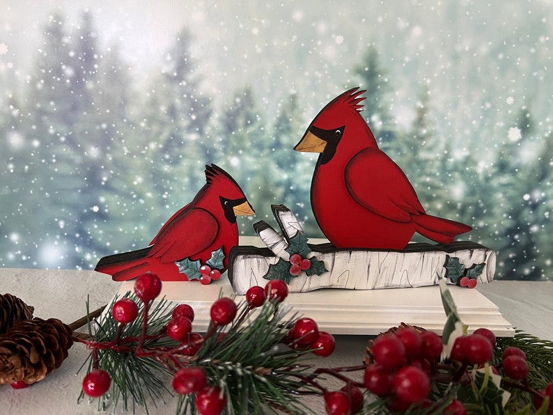 Pode incluir: Uma decora&ccedil;&atilde;o de Natal de madeira com dois cardeais vermelhos empoleirados em um galho de b&eacute;tula branco com azevinho verde e bagas vermelhas. Os cardeais s&atilde;o pintados em um estilo realista com olhos e bicos pretos. O fundo &eacute; uma imagem desfocada de uma floresta nevada.