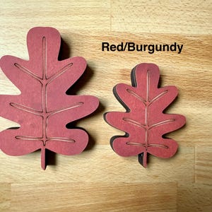 May include: Two red and burgundy oak leaf-shaped cutouts of different sizes. Each leaf features a central stem and branching veins etched into the surface. The text Red/Burgundy is printed in black above the smaller leaf on the light wood surface.