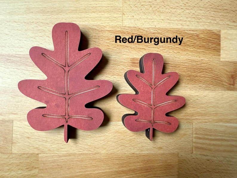 May include: Two red and burgundy oak leaf-shaped cutouts of different sizes. Each leaf features a central stem and branching veins etched into the surface. The text Red/Burgundy is printed in black above the smaller leaf on the light wood surface.