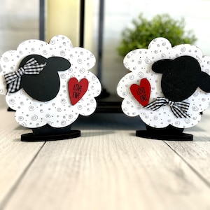 May include: Two wooden sheep figurines with black and white swirl patterns. Each sheep has a black head and a red heart with the text "Love Ewe" on it. The sheep are standing on black bases.
