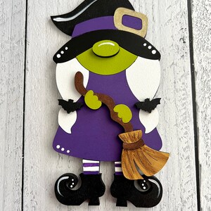 May include: A purple and green witch gnome with a black hat and a brown broom. The gnome is wearing white and purple striped socks and black shoes.