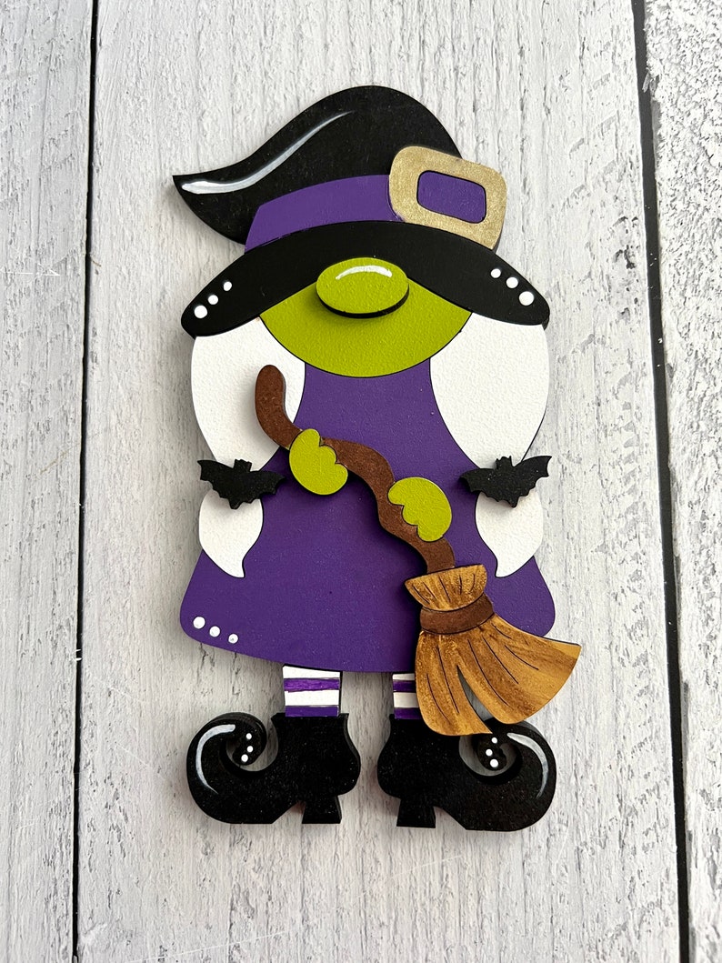 May include: A purple and green witch gnome with a black hat and a brown broom. The gnome is wearing white and purple striped socks and black shoes.