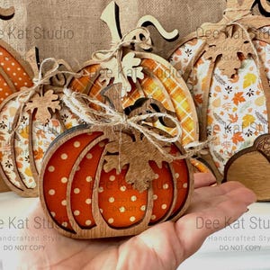 May include: A collection of layered wooden pumpkin decorations featuring various patterns including orange polka dots, autumn leaves, and plaid. Each pumpkin is accented with a small wooden leaf cutout and tied with a natural jute twine bow.