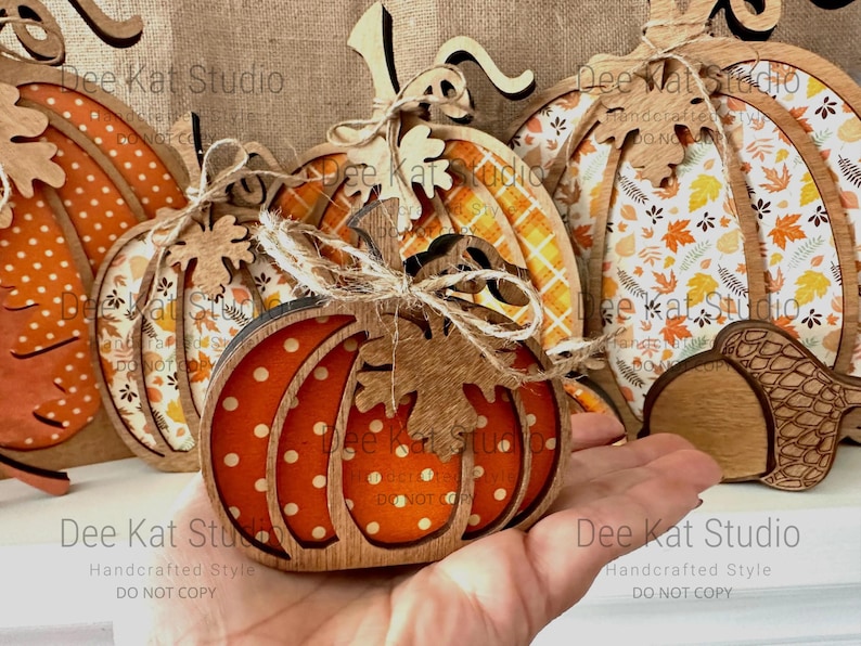 May include: A collection of layered wooden pumpkin decorations featuring various patterns including orange polka dots, autumn leaves, and plaid. Each pumpkin is accented with a small wooden leaf cutout and tied with a natural jute twine bow.