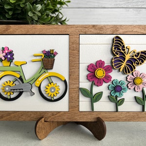 K&ouml;nnte beinhalten: Ein Wandbild in einem Holzrahmen mit zwei separaten Abschnitten. Der linke Abschnitt zeigt ein gelbes Fahrrad mit einem Blumenkorb. Der rechte Abschnitt zeigt einen lila Schmetterling und drei Blumen in rosa, hellrosa und blau.