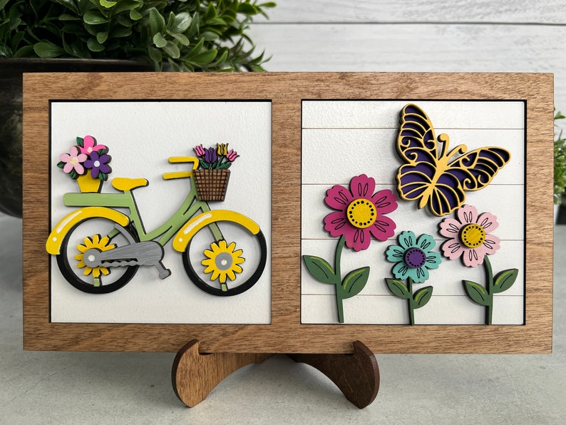 K&ouml;nnte beinhalten: Ein Wandbild in einem Holzrahmen mit zwei separaten Abschnitten. Der linke Abschnitt zeigt ein gelbes Fahrrad mit einem Blumenkorb. Der rechte Abschnitt zeigt einen lila Schmetterling und drei Blumen in rosa, hellrosa und blau.