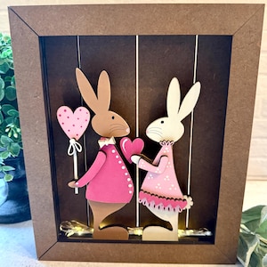 May include: A wooden framed art piece featuring two rabbit figures. One rabbit holds a pink heart-shaped balloon, the other holds a pink heart. Both rabbits wear pink outfits. The background is a dark brown with vertical lines.