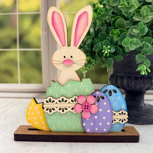 May include: Wooden Easter decoration featuring a bunny emerging from a cracked egg, with three colorful eggs. The bunny has pink-lined ears and a pink nose. The eggs are yellow, green, purple, and blue, with polka dots and floral accents. The base is brown.