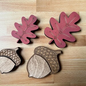 May include: Two wooden oak leaves painted in a deep red with etched vein details and two wooden acorns featuring natural light wood grain on the nut portion and dark etched cross-hatch patterns on the caps, arranged in two distinct sizes.