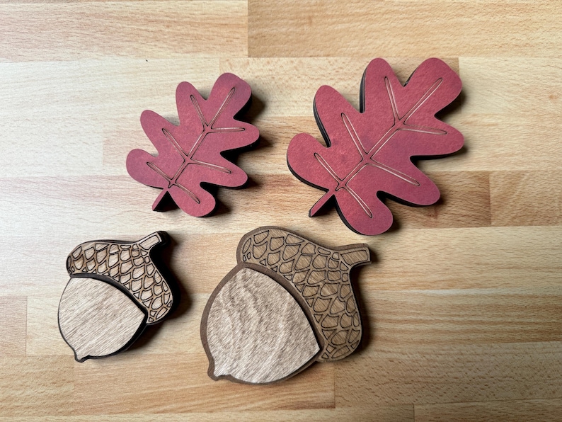 May include: Two wooden oak leaves painted in a deep red with etched vein details and two wooden acorns featuring natural light wood grain on the nut portion and dark etched cross-hatch patterns on the caps, arranged in two distinct sizes.