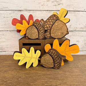 May include: A collection of wooden autumn-themed decorations featuring three brown acorns with textured caps and four flat leaves in shades of red, orange, and yellow. The items are arranged in front of two small, dark brown wooden crates.