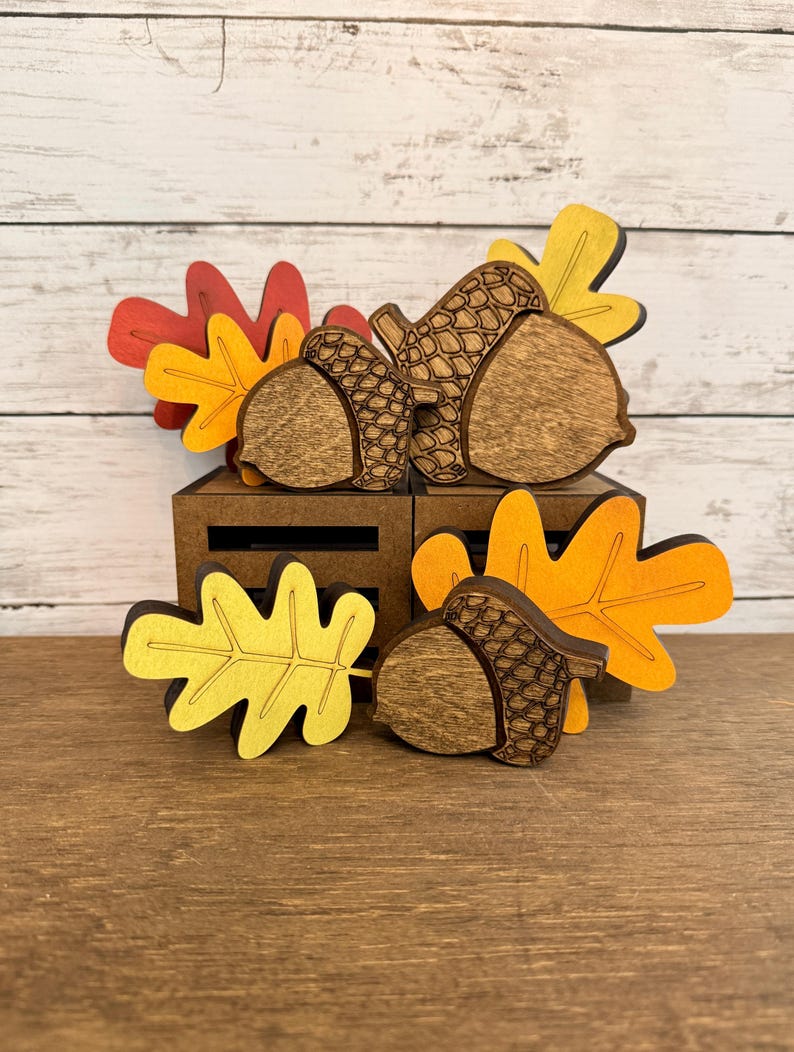 May include: A collection of wooden autumn-themed decorations featuring three brown acorns with textured caps and four flat leaves in shades of red, orange, and yellow. The items are arranged in front of two small, dark brown wooden crates.