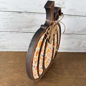 May include: A pumpkin-shaped wooden decoration with a dark brown stained edge and a central panel featuring a pattern of orange, yellow, and brown autumn leaves. A small wooden leaf cutout is tied to the stem with a thin piece of twine.