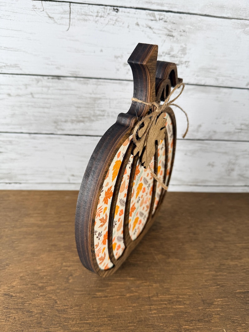 May include: A pumpkin-shaped wooden decoration with a dark brown stained edge and a central panel featuring a pattern of orange, yellow, and brown autumn leaves. A small wooden leaf cutout is tied to the stem with a thin piece of twine.