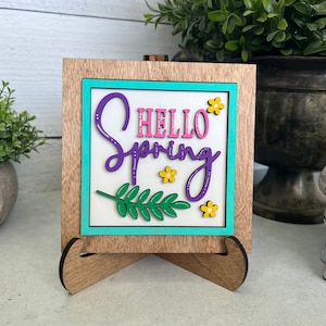 K&ouml;nnte beinhalten: Ein Holzschild mit einem t&uuml;rkisfarbenen Rahmen und wei&szlig;em Hintergrund. Das Schild sagt "Hello Spring" in lila und rosa Schrift mit einem gr&uuml;nen Blatt und gelben Blumen.