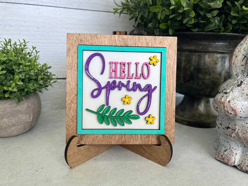 K&ouml;nnte beinhalten: Ein Holzschild mit einem t&uuml;rkisfarbenen Rahmen und wei&szlig;em Hintergrund. Das Schild sagt "Hello Spring" in lila und rosa Schrift mit einem gr&uuml;nen Blatt und gelben Blumen.