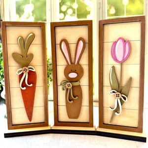 May include: Three wooden Easter decorations. The first is a carrot with a green top, the second is a brown bunny with pink ears, and the third is a pink tulip. Each is framed in a brown wooden frame.