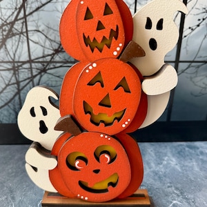 May include: A Halloween-themed wooden decoration featuring three stacked orange jack-o'-lanterns with carved faces and a white ghost. The pumpkins have brown stems and are accented with white dots. The decoration sits on a wooden base.