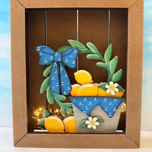 May include: Wooden framed art piece featuring a lemon-themed design. The artwork includes a blue bow with white polka dots, lemons, and a basket with a blue polka dot cloth. The frame is brown, and the background has vertical lines.