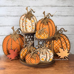 May include: A tiered wooden display stand holding multiple flat pumpkin-shaped decorations. The pumpkins feature various orange and white patterns including polka dots, plaid, and autumn leaves. Each pumpkin is topped with a wooden leaf cutout.