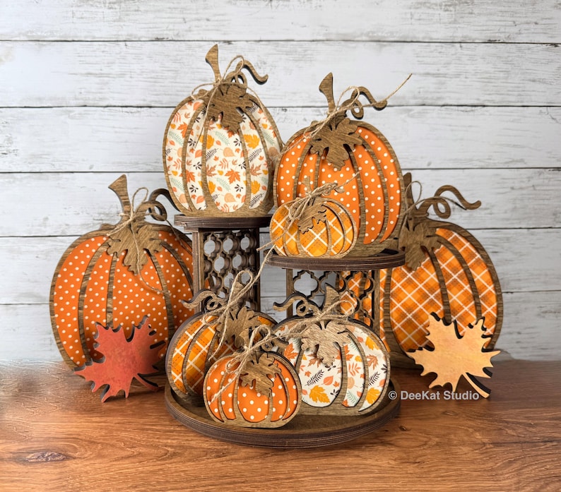 May include: A tiered wooden display stand holding multiple flat pumpkin-shaped decorations. The pumpkins feature various orange and white patterns including polka dots, plaid, and autumn leaves. Each pumpkin is topped with a wooden leaf cutout.