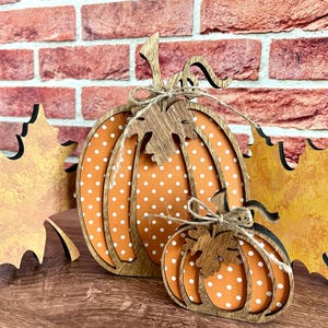 May include: Two decorative wooden pumpkins featuring an orange surface with white polka dots and dark brown wood-toned outlines. Each pumpkin is adorned with a wooden leaf cutout and a tied piece of natural jute twine at the top stem.