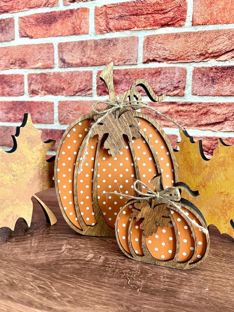 May include: Two decorative wooden pumpkins featuring an orange surface with white polka dots and dark brown wood-toned outlines. Each pumpkin is adorned with a wooden leaf cutout and a tied piece of natural jute twine at the top stem.