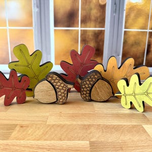 May include: A collection of wooden autumn-themed decorations featuring four oak leaves in shades of red, yellow, and green, alongside two acorns with natural wood grain bodies and dark etched caps, all standing upright on a light wood surface.