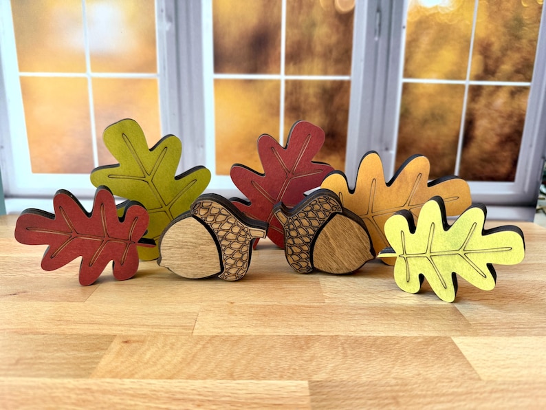 May include: A collection of wooden autumn-themed decorations featuring four oak leaves in shades of red, yellow, and green, alongside two acorns with natural wood grain bodies and dark etched caps, all standing upright on a light wood surface.