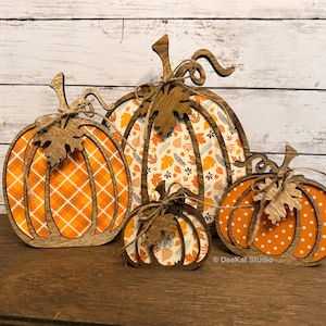 May include: Four decorative wooden pumpkin cutouts of varying sizes. Each pumpkin features a dark wood frame with a patterned orange and white insert, a wooden leaf accent, and a tied twine bow. The text DeeKat Studio is visible at the bottom.