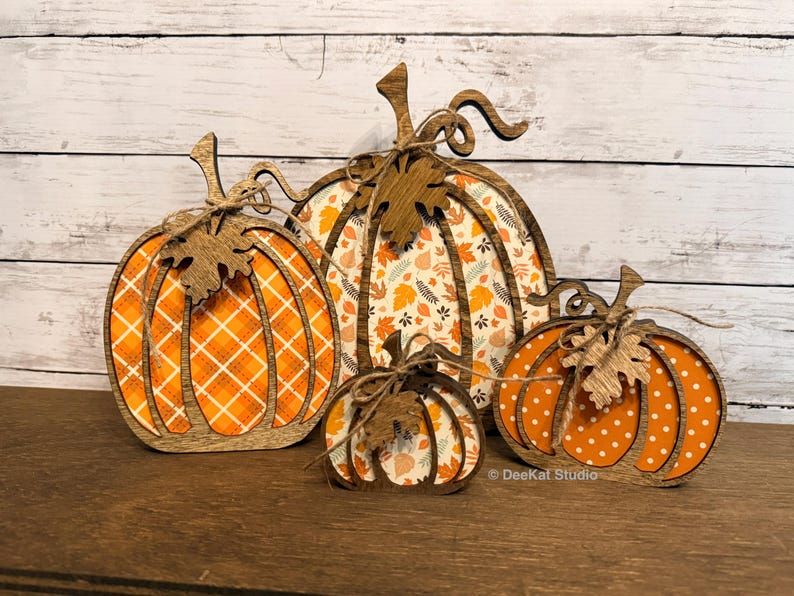 May include: Four decorative wooden pumpkin cutouts of varying sizes. Each pumpkin features a dark wood frame with a patterned orange and white insert, a wooden leaf accent, and a tied twine bow. The text DeeKat Studio is visible at the bottom.