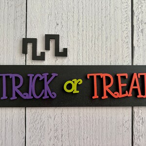 May include: Black sign with the words "Trick or Treat" in purple, green, and orange letters. The sign is on a white wood background.