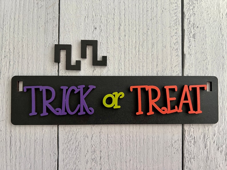 May include: Black sign with the words "Trick or Treat" in purple, green, and orange letters. The sign is on a white wood background.