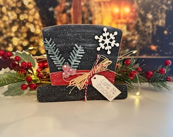 May include: A black wooden snowman hat with a red ribbon, decorative greenery, and a tag that reads "Let it snow." The hat is decorated with a white snowflake and sprigs of greenery. Festive holiday decor.
