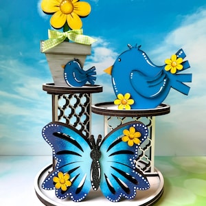 May include: A decorative wooden display featuring a blue butterfly, two blue birds, and a flower in a pot. The butterfly and birds are adorned with yellow flowers. The display is set against a blue sky backdrop.