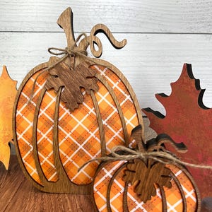 May include: Two wooden pumpkin-shaped decorations featuring a cutout frame design with an orange and white plaid pattern background. Each pumpkin is accented with a dark brown wooden leaf cutout and a tied piece of thin brown twine.